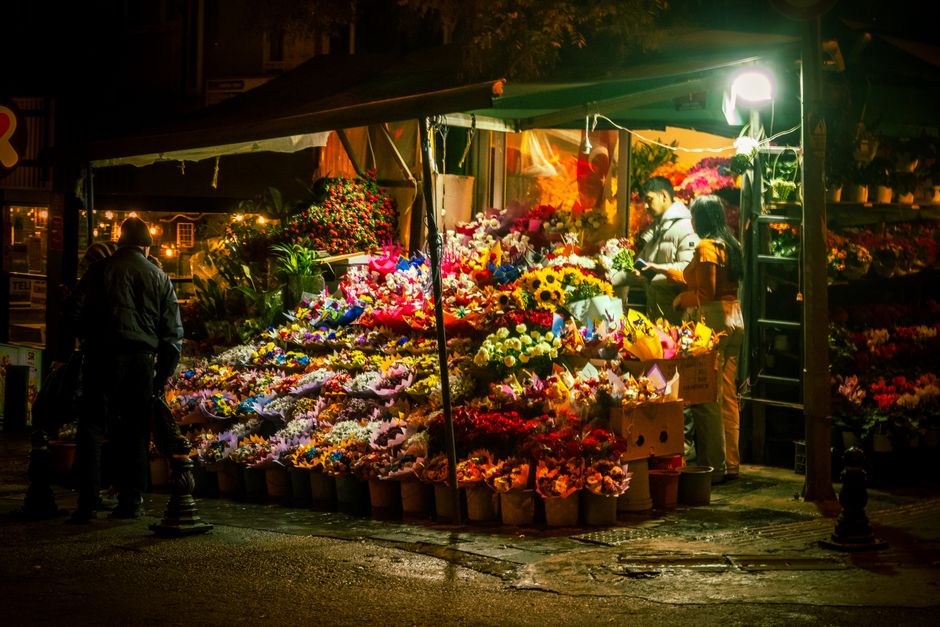 A vibrant flower stall illuminated by bright lighting at night, showcasing an extensive display of fresh floral arrangements. The bouquet arrangements include sunflowers, roses, tulips, lilies, and da