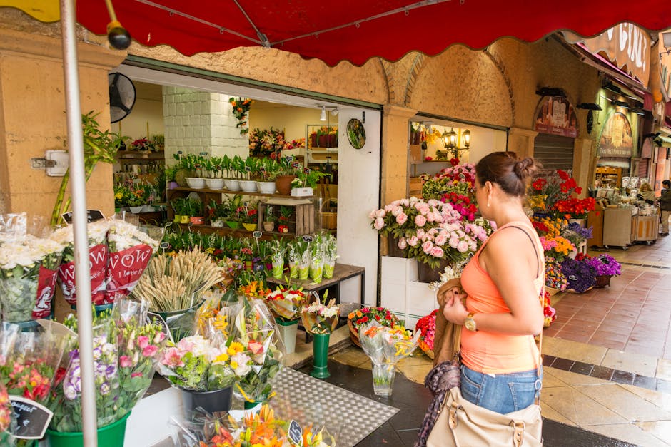 A woman in an orange sleeveless top and blue jeans stands outside a flower shop under a red awning, holding a beige handbag and a brown jacket, gazing at a display of fresh floral arrangements. The sh