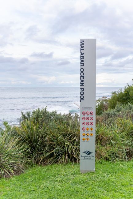 A tall, rectangular white sign with black text reading 'Malabar Ocean Pool' stands amidst green shrubbery and coastal vegetation, overlooking the ocean with a partly cloudy sky overhead. The sign incl