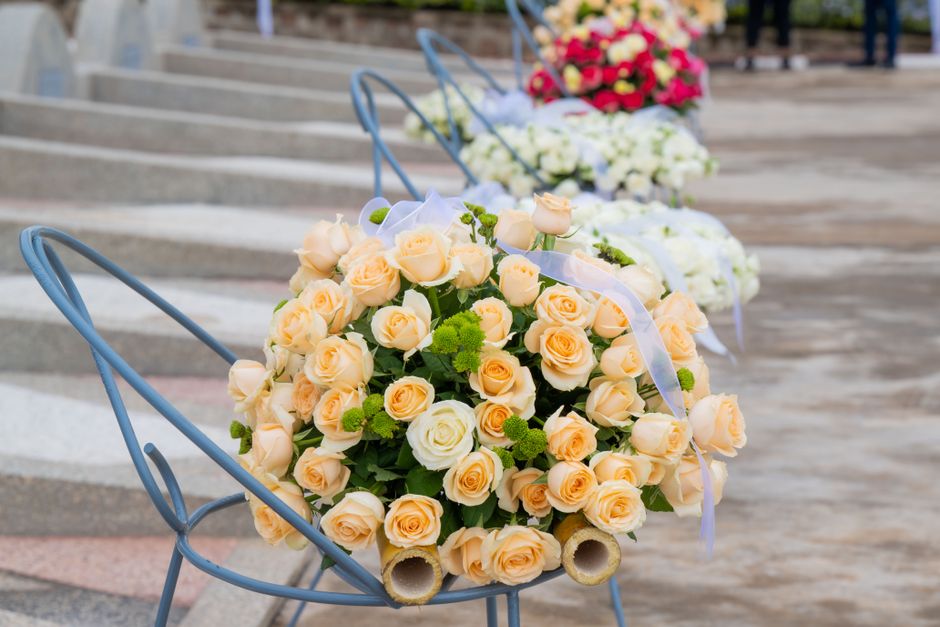 A floral arrangement featuring a bouquet of pale peach and ivory roses with small green pom-pom-like accents, displayed in a metal cart with a curved handle, positioned outdoors on a paved surface wit