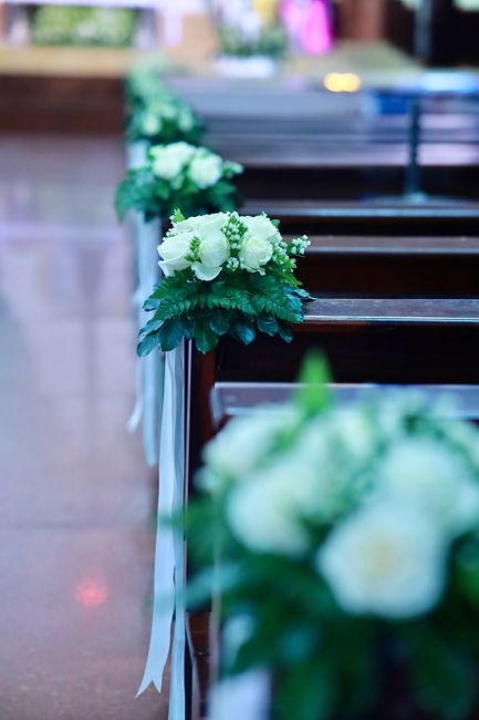 A close-up view of a wedding floral arrangement featuring a small bouquet of white roses, ranunculus, and green foliage with delicate white ribbons, placed atop a polished wooden pew at St Johns Gate.